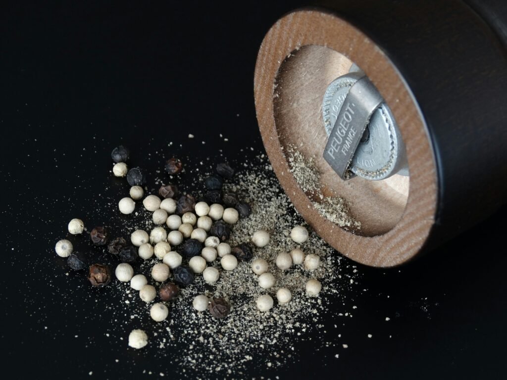 Close-up of a pepper grinder and assorted peppercorns on black surface.