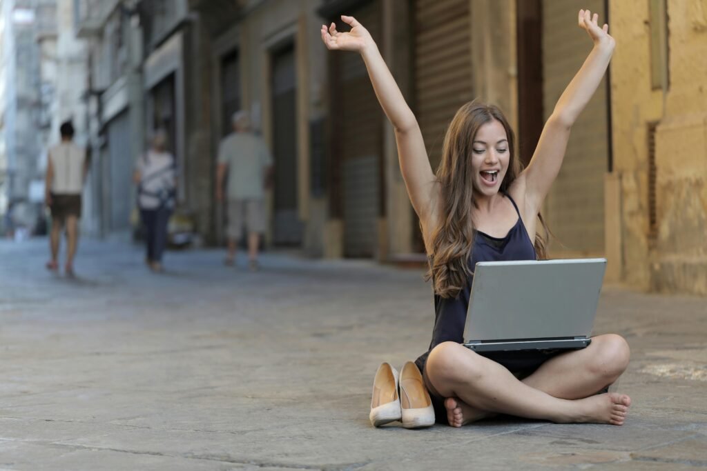 A cheerful woman sitting outdoors, celebrating success with arms raised, while using a laptop.