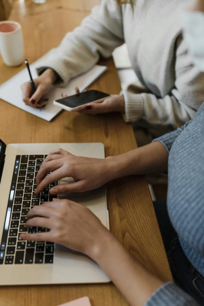 Two people collaborating in workspace with laptop and smartphone, typing notes.